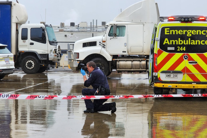 A police officer kneeling on a wet road holds a camera up to his eye. In the background are white trucks and an ambulance