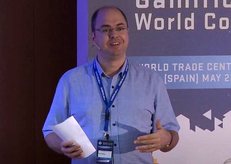 A balding man wearing a headset, blue shirt, a lanyard around his neck.