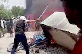 A man surrounded by a mob hits a piece of furniture on fire with a stick. 