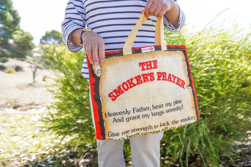 A lady's hands hold a cream and red canvas tote bag with the words 'the smoker's prayer' on it.