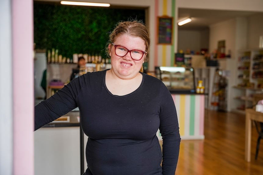 A young woman wearing glasses and a black top holds open a cafe door, smiling. Ausnew Home Care, NDIS registered provider, My Aged Care