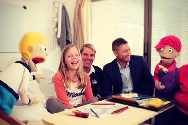 Young girl sits on a bed smiling next to two puppets and two men