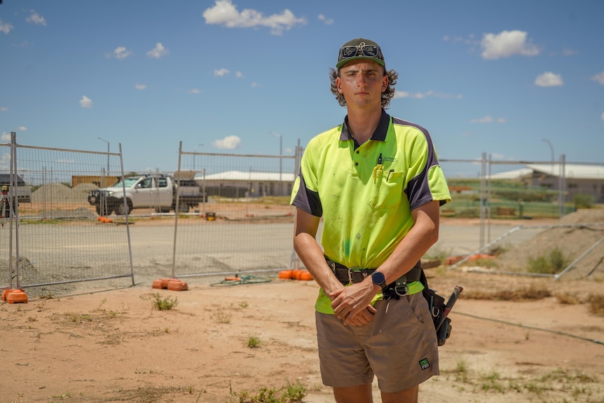 A young man wearing a flat-cap and high-vis workwear stands in a home construction site on a sunny day.