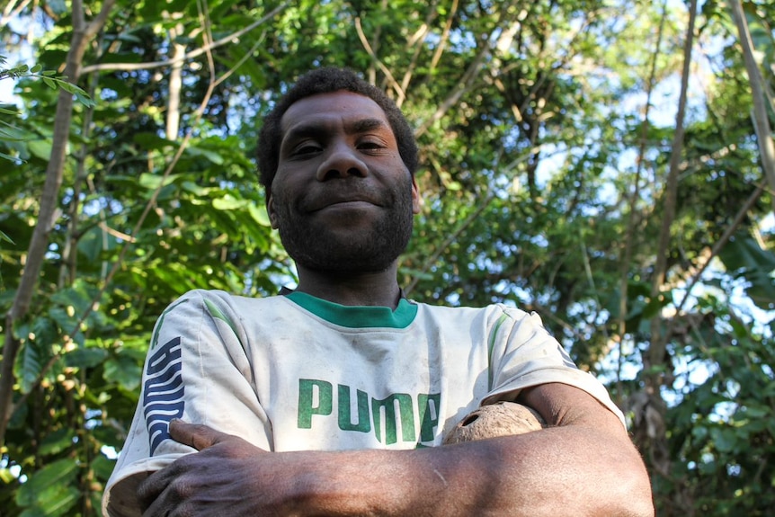A man from Vanuatu folds his arms as he poses for a photo, with trees behind him.