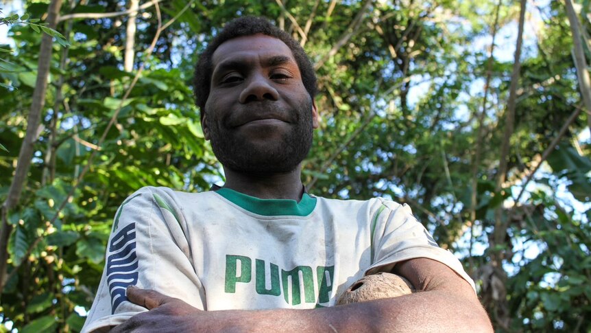 A man from Vanuatu folds his arms as he poses for a photo, with trees behind him.