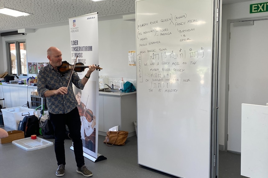 A man playing a violin in a classroom with a whiteboard with song lyrics.