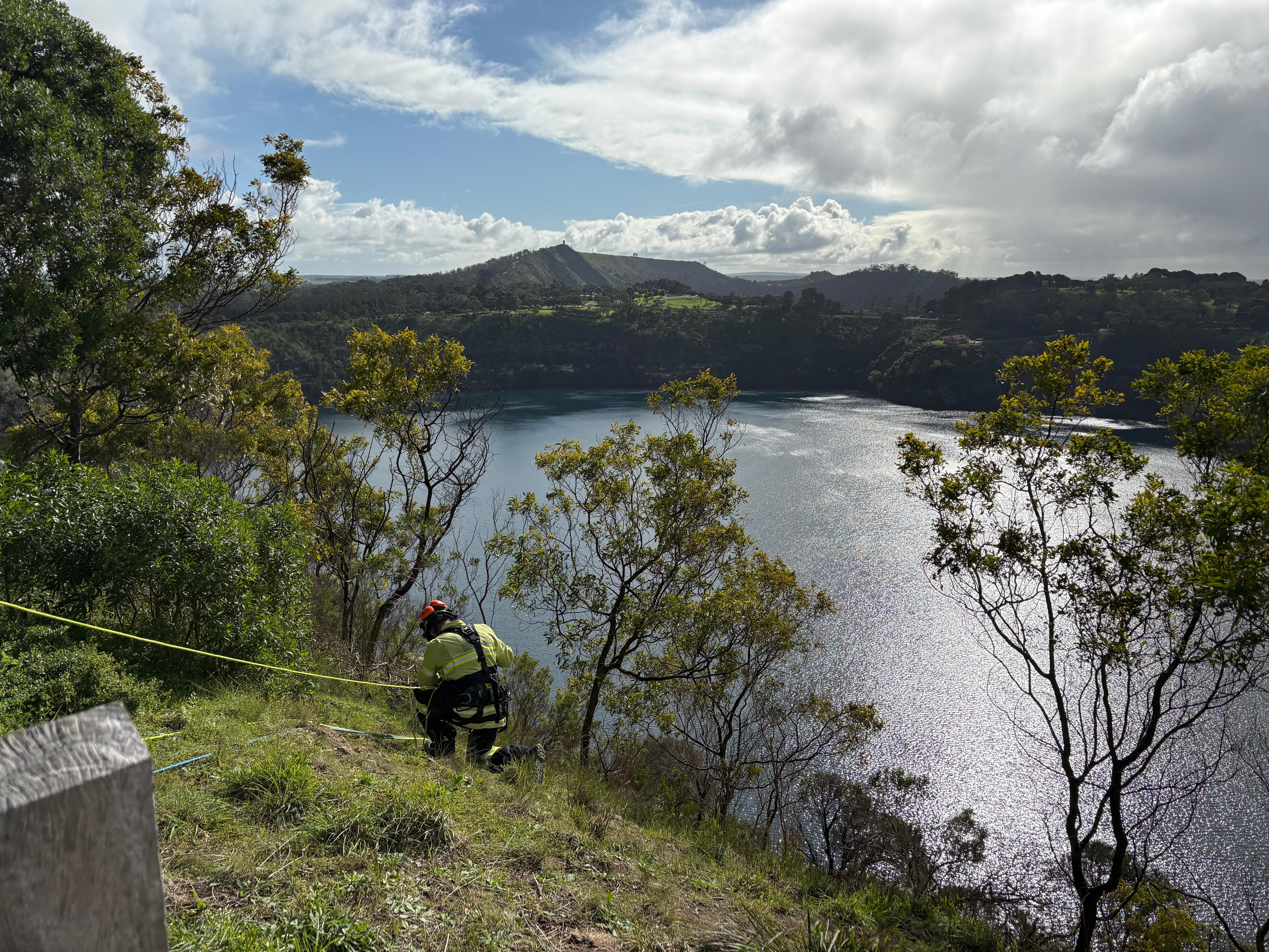 a man attached to a rope kneeling on a cliff, with a lake below