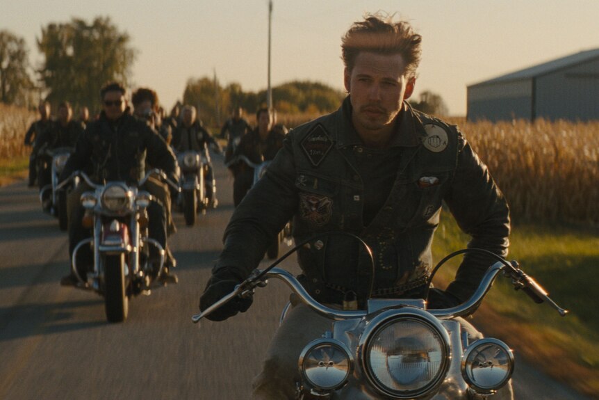 Austin Butler riding a motorbike on a road through a field, with others riding behind him