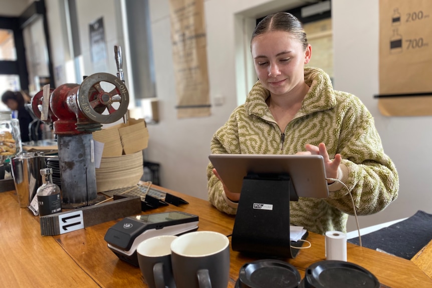 A smiling young woman operates an iPad behind the counter of a cafe.