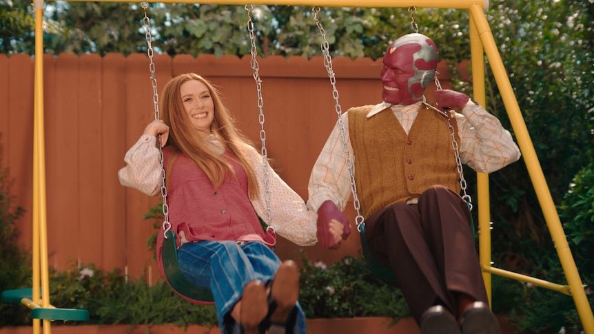 A 30-something woman and a masked man in 70s dress hold hands while smiling broadly and swinging on a yellow backyard swing set.