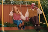 A 30-something woman and a masked man in 70s dress hold hands while smiling broadly and swinging on a yellow backyard swing set.