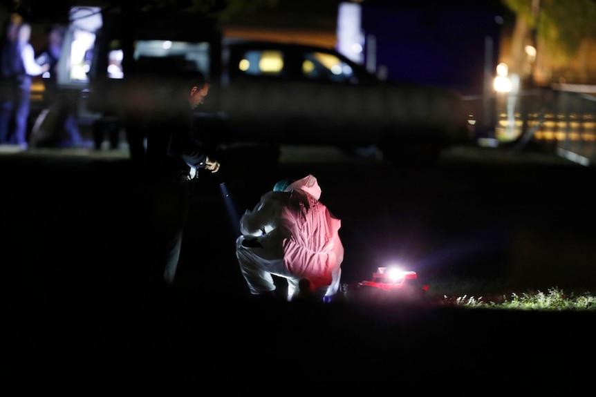 An officer in a red forensic suit squats down in the park at night with a torch.