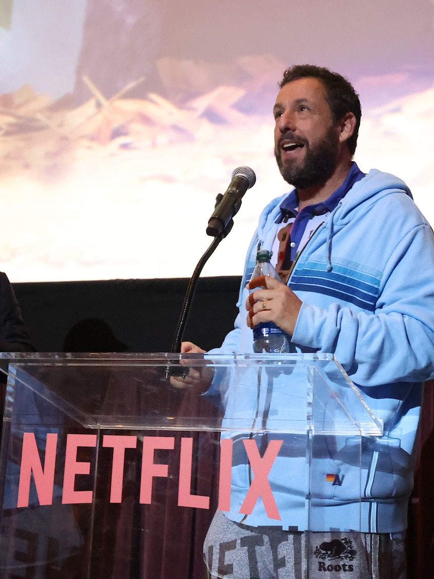 Adam Sandler holds the microphone, standing at the Netflix podium at a cinema screening of Leo.