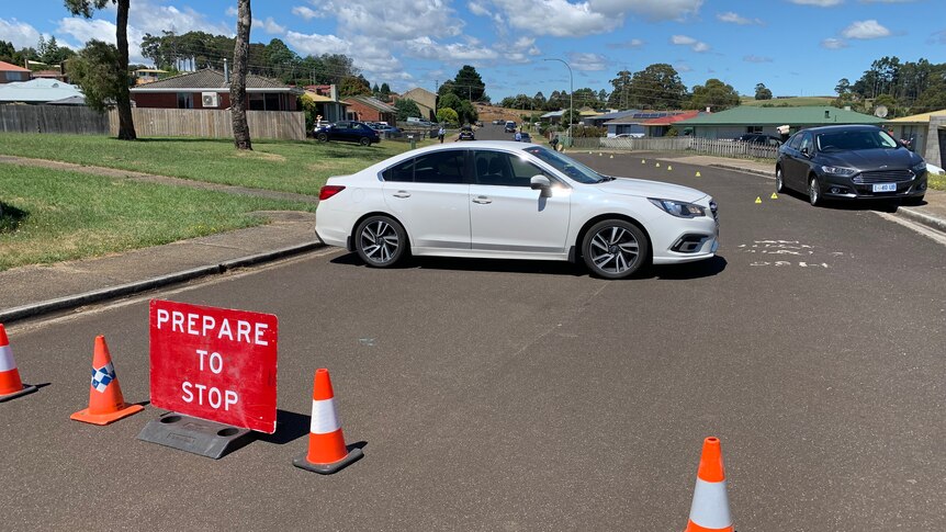 A white car in the middle of the road behind a sign that reads "prepare to stop". Crime scene markings in the background.