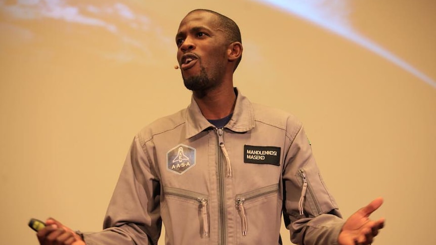 An African man gesticulates in a grey space jumpsuit in front of a sand-coloured screen with a close-up of the earth.