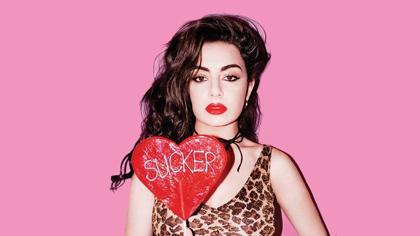 Charli XCX in a leopard print top, in front of a pink background, holding a huge love heart lollypop.