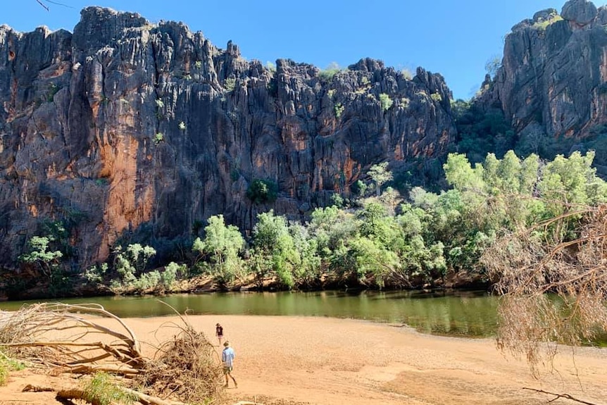 Two people in the distance, on sand with a large cliff in the distance and a waterhole in front