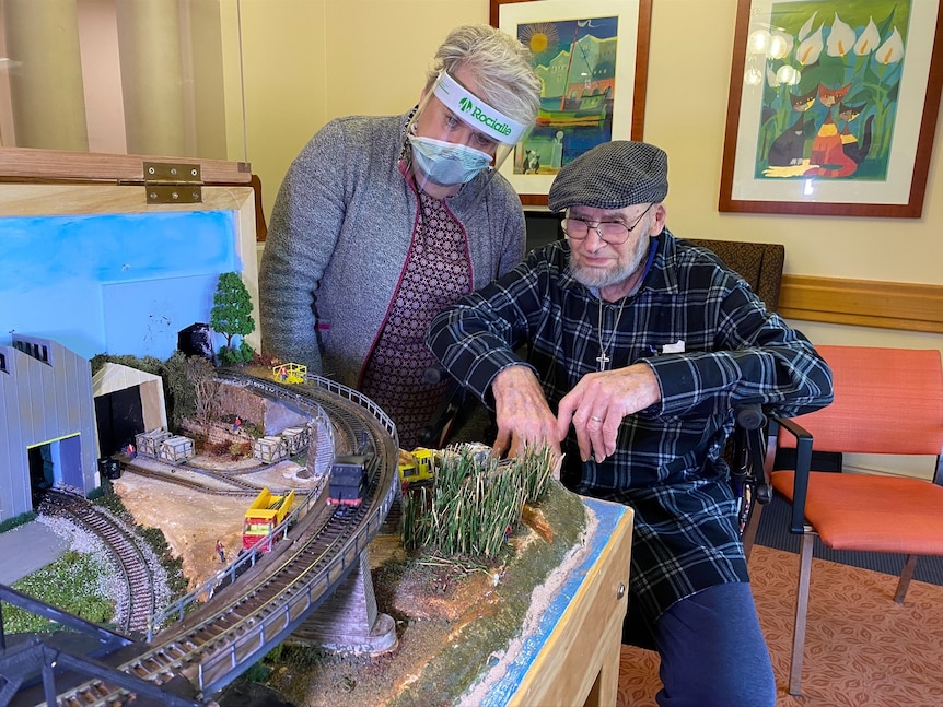 Man with grey beard and female carer beside model train set