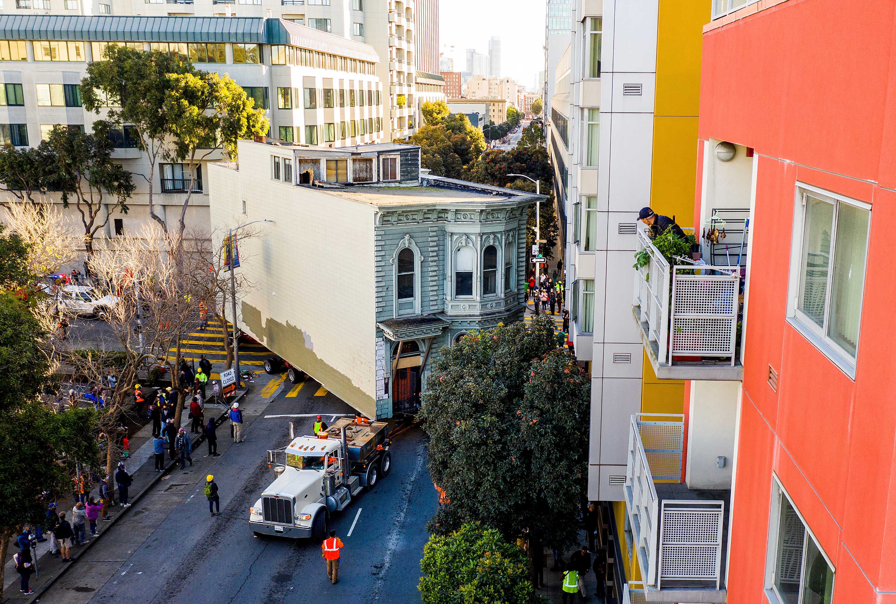 A man stands on a balcony on the right as he watches a truck pulling a house through the street below him.