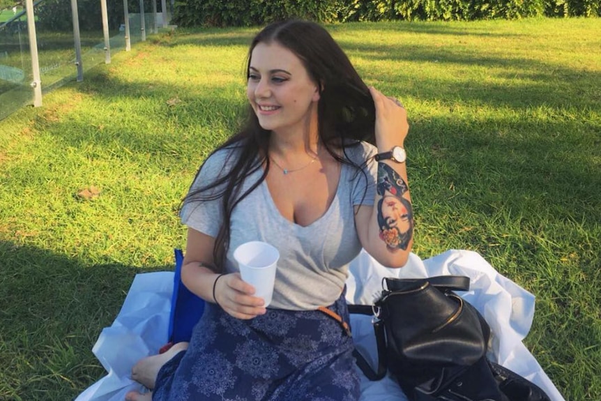 A young woman sitting on a blanket in a park smiling and holding a drink.