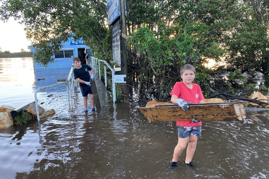 Young boys Finley and Alastair O'Donnell wade through water to help to clean up flood damage at Big Oyster in Port Macquarie.