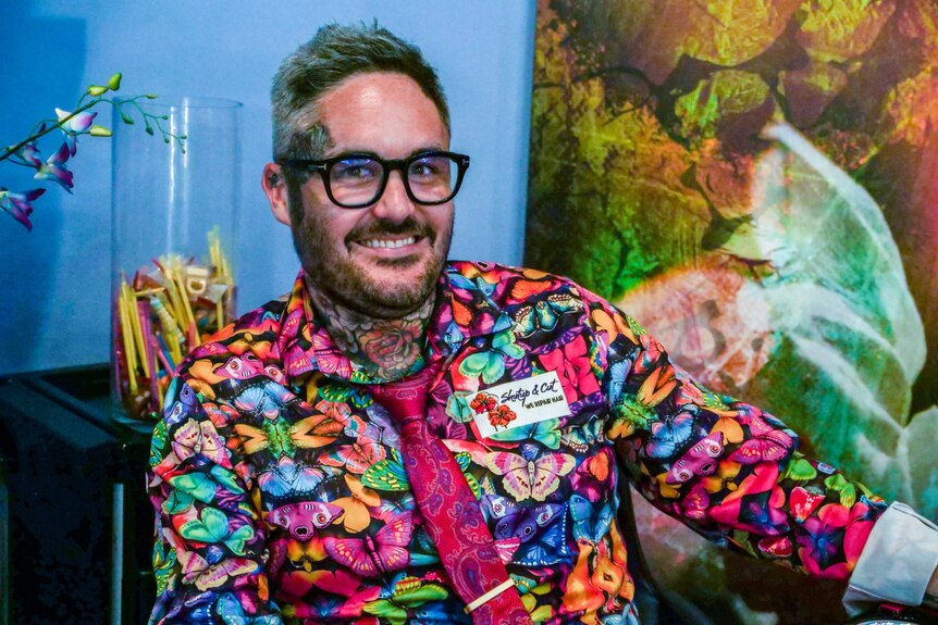 A male hairdresser wearing a colourful butterfly shirt sits in front of a bowl of lollies and artwork in his salon.
