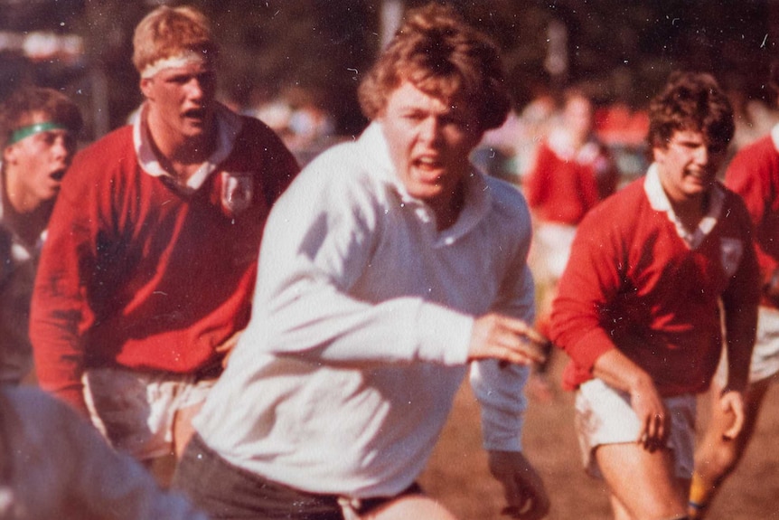 A football player, surrounded by other men, runs to chase the player with the ball.