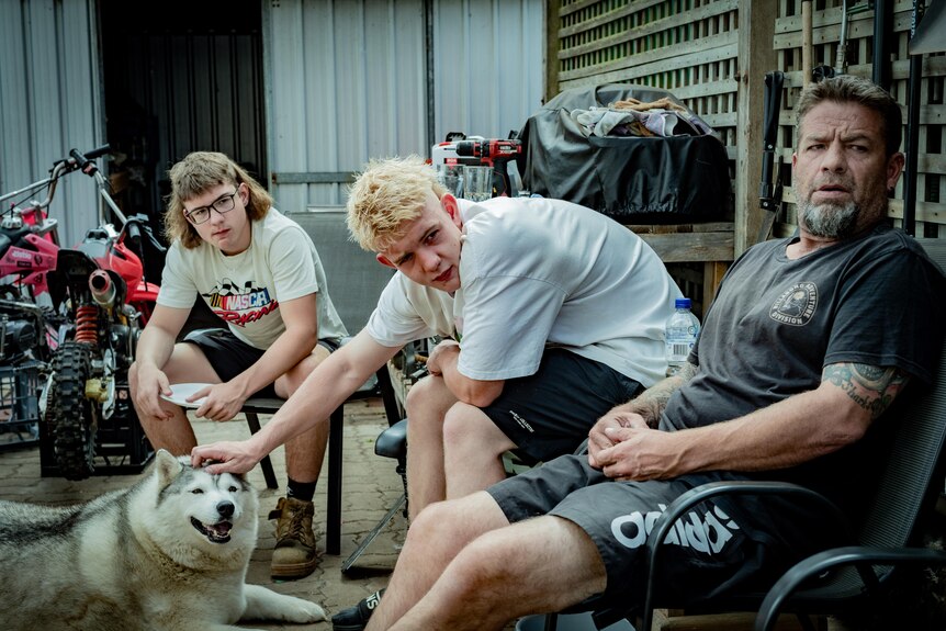 Two teenage boys and their dad sit in the backyard with their husky. 