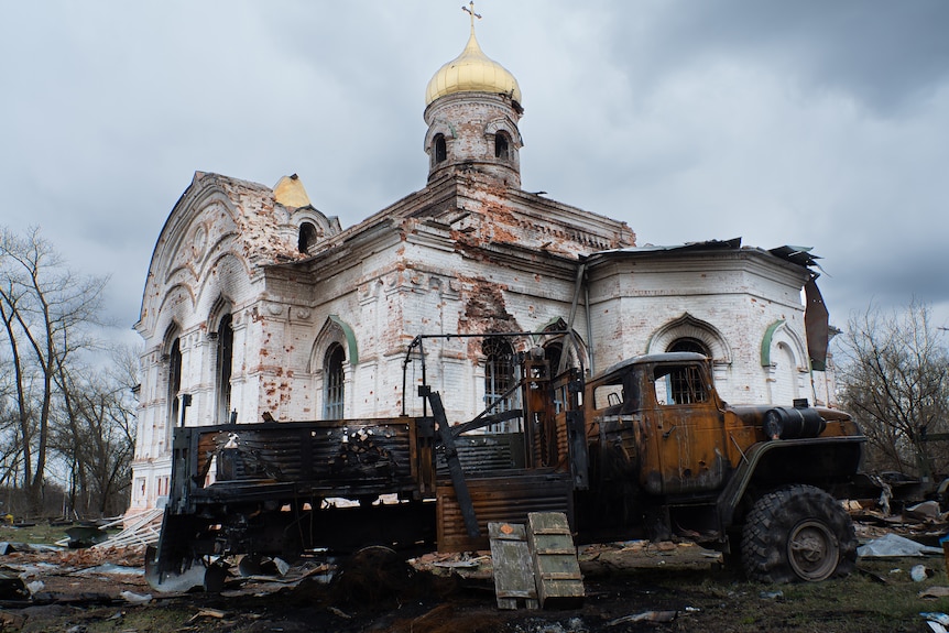 A burned out church with a golden spire, with the shell of a destroyed armoured vehicle out the front
