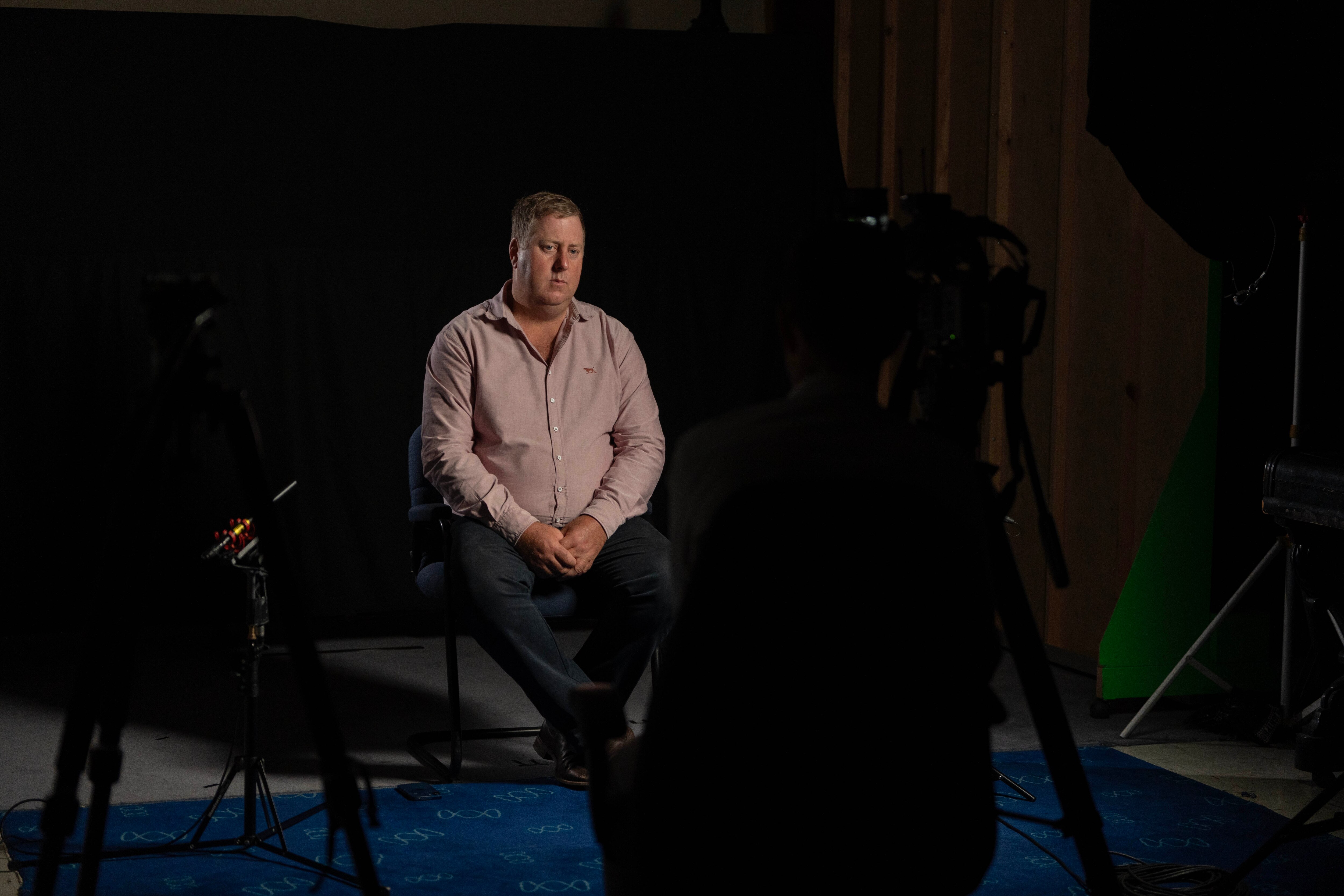 Un hombre con una camisa de cuello rosa se sienta en una habitación oscura durante una entrevista.