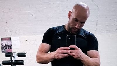 Stavro D'Amore wearing a tight black shirt with muscles exposed, wearing grey pants, standing in a gym taking a photo of himself