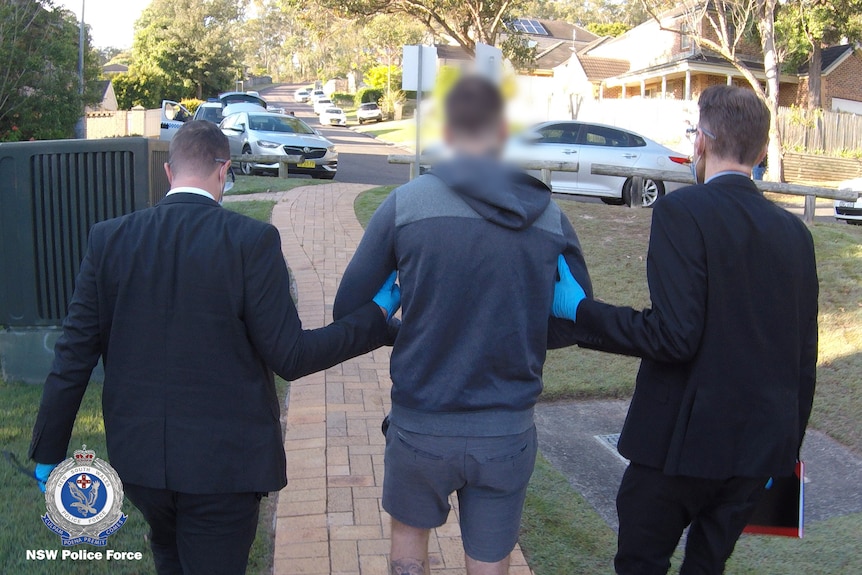 Two detectives walk alongside a man in hand-cuffs, accused of fraud and drug supply.