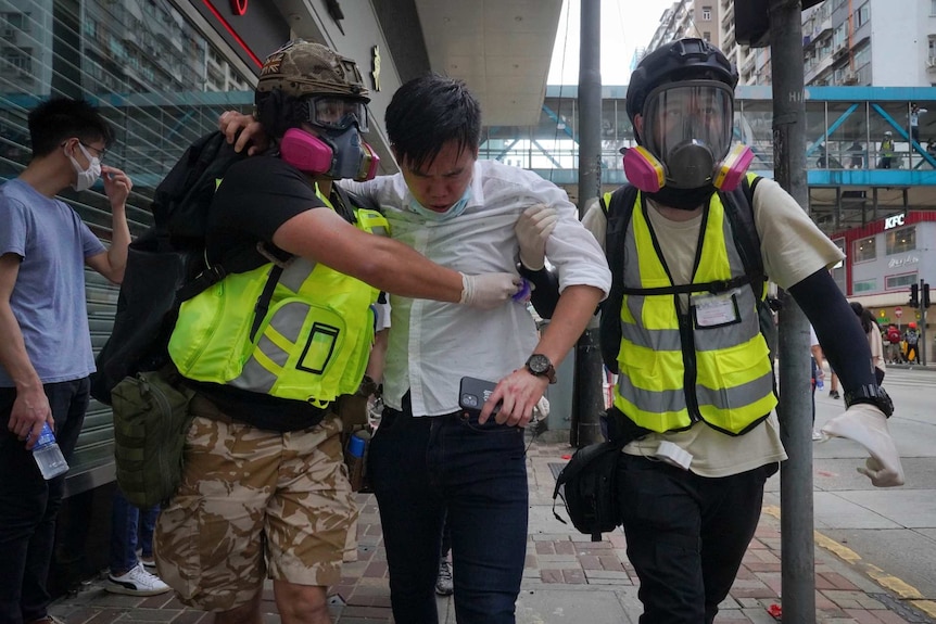 Women in face masks and florescent vests lead a man in a white shirt with liquid on his face away