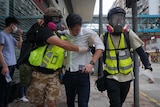 two men in face masks and florescent vests lead a man in a white shirt with liquid on his face away