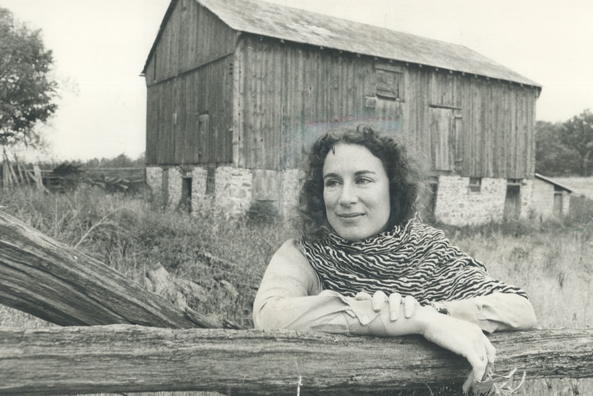 A black and white photo of a young woman leaning on a fence, standing in front of a barn