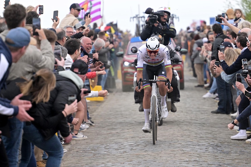 A professional cyclist on cobble stone roads, riding through a cheering crowd as he leads the Paris-Roubaix race