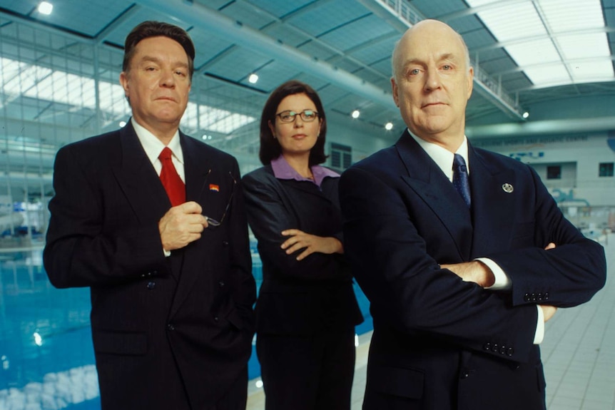 Bryan Dawe, Gina Riley and John Clarke stand by an Olympic swimming pool.