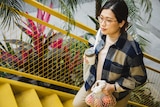 A young woman with black hair and glasses walks up interior stairs holding cloth shopping bag full of apples and a reusable cup.