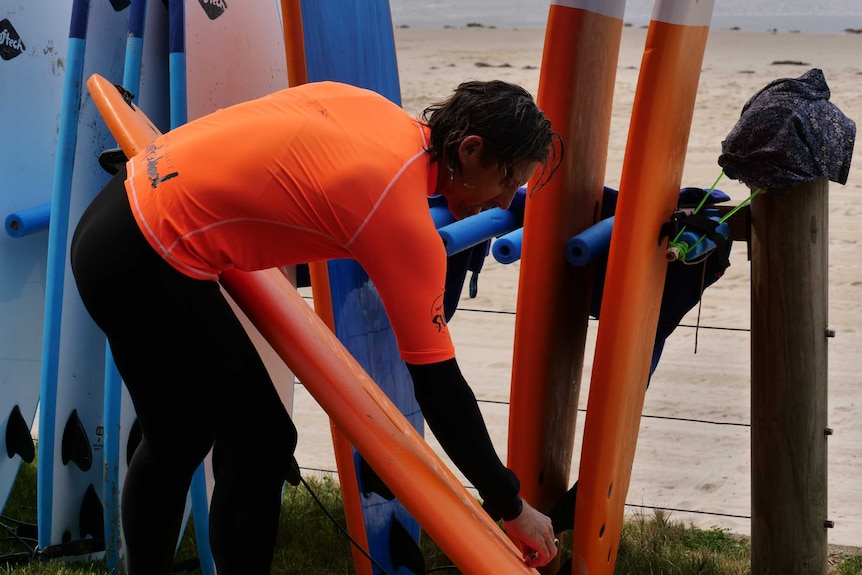 A surfer in a wetsuit leaning over waxing a surfboard by the beach.