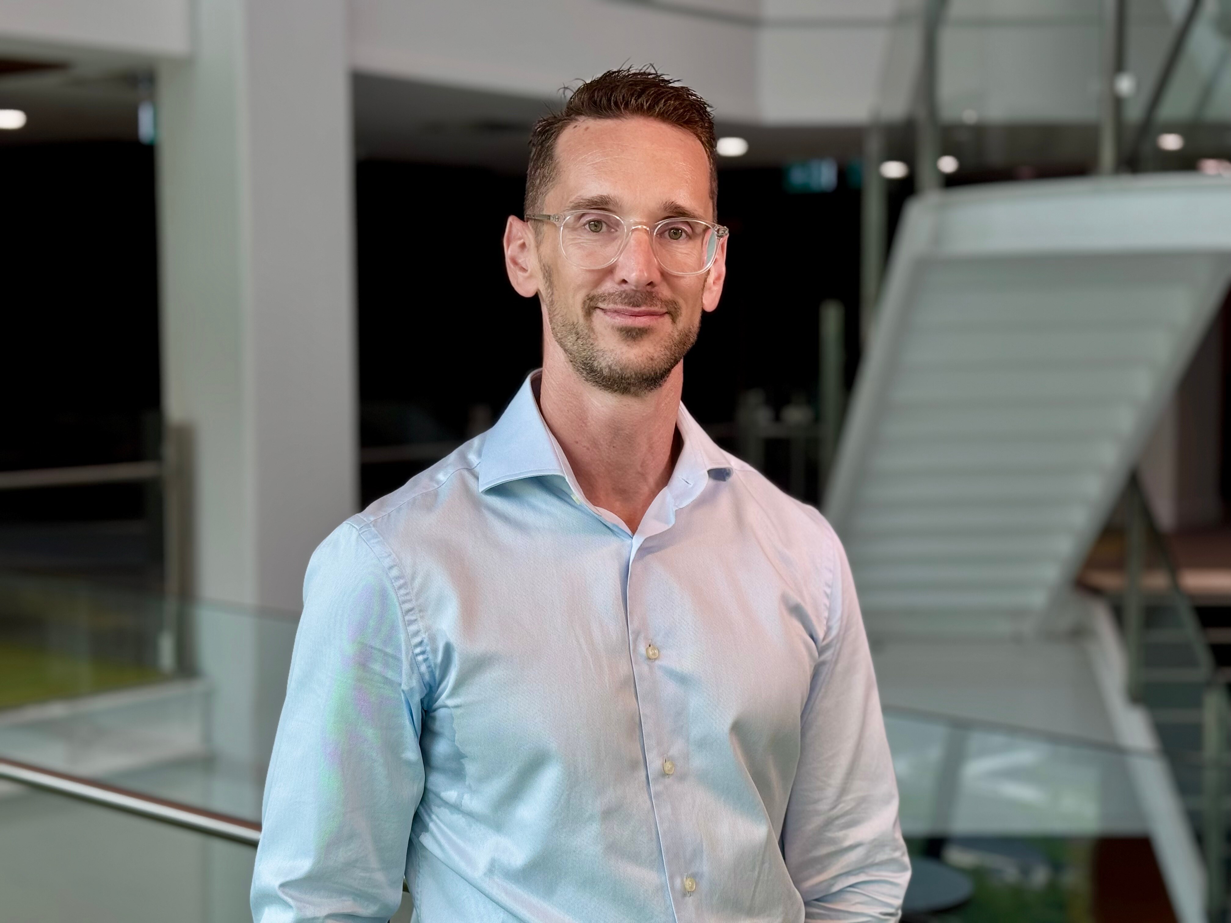 Bespectacled man wearing light blue shirt standing in a foyer with stairs in the background