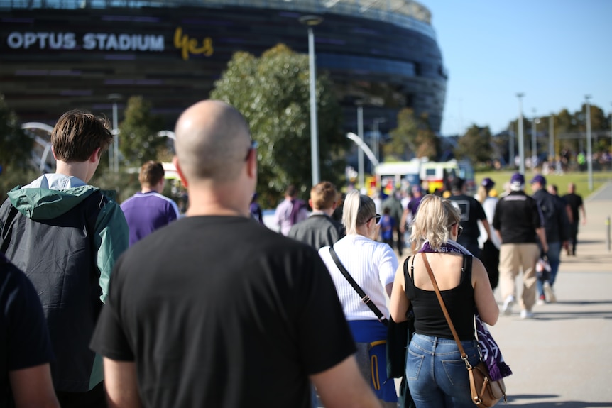 Crowds are walking along a footpath leading to Perth Stadium on a sunny day wearing Dockers scarves among other football merch
