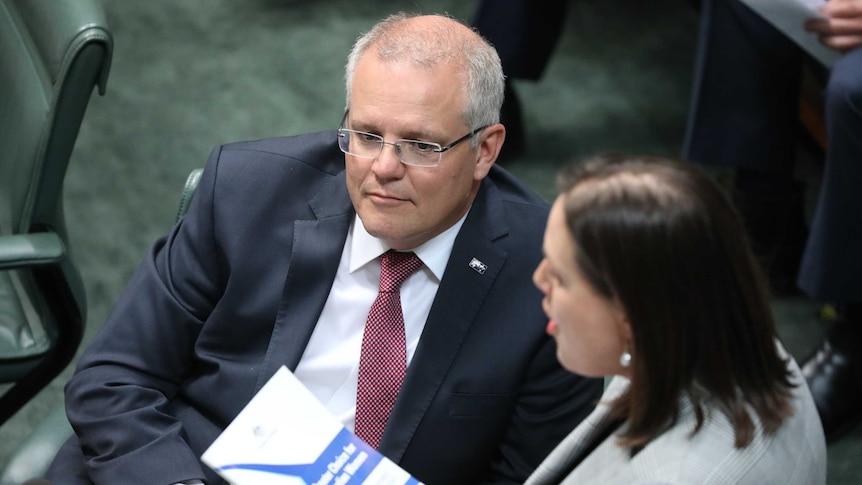 a man with glasses in a suit sitting in parliament watches as a woman stands and speaks