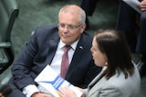 a man with glasses in a suit sitting in parliament watches as a woman stands and speaks