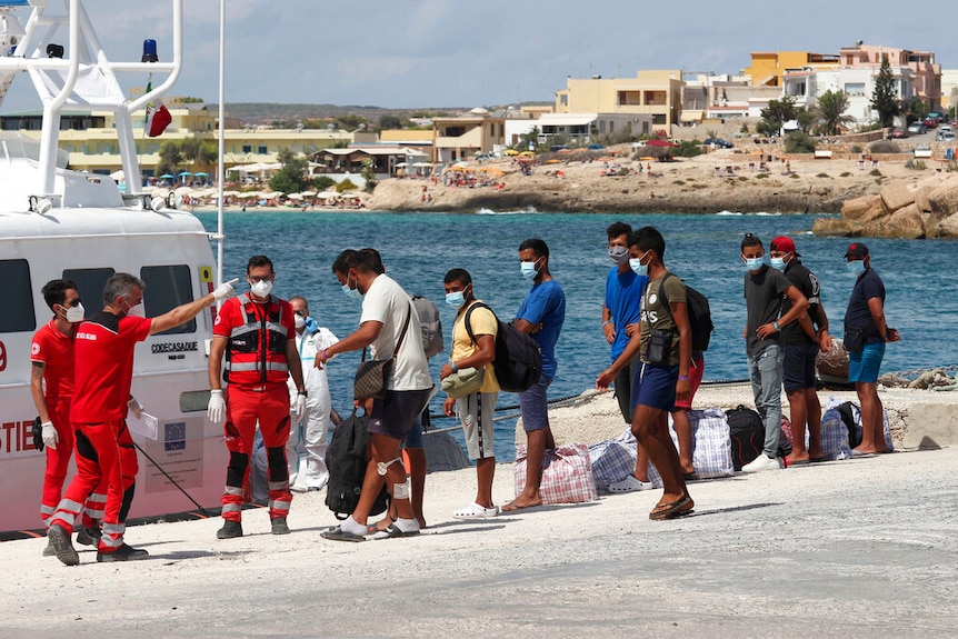 On a bright day, you view a line of male asylum seekers of next to emergency workers in red at a jetty.
