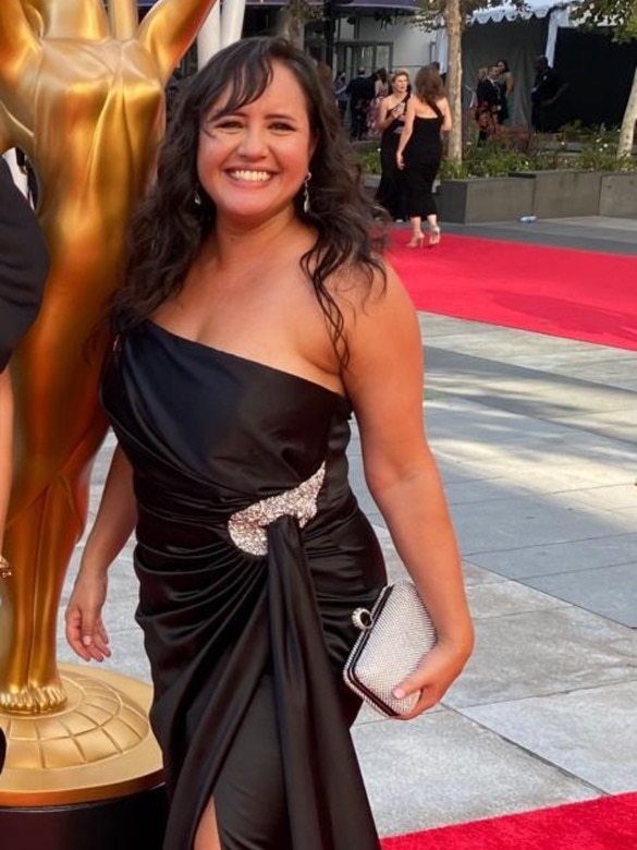 A woman in a ballgown poses with a smile in front of a golden statue on a red carpet.