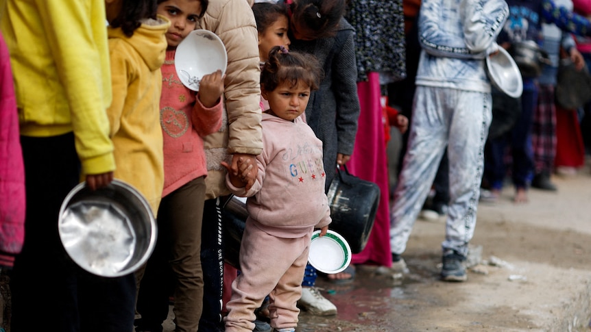 A little girl, holding an empty white bowl, frowns as she lines up with others who are also holding empty food containers