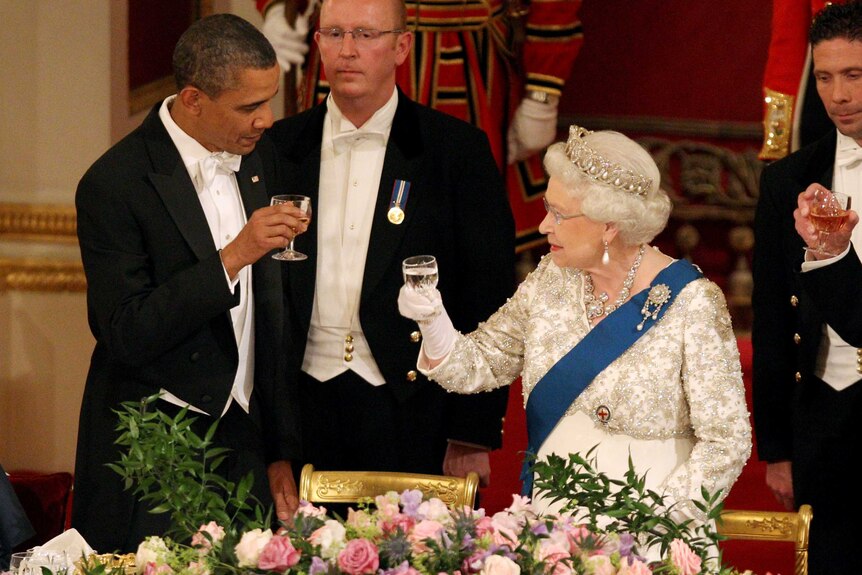 Barack Obama - dressed in tuxedo - and the Queen - in a jewel-adorned dress - look at each other, holding glasses in a toast.