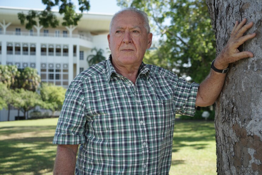 A elderly man wearing a checked shirt leans his hand against a tree and looks into the distance. 