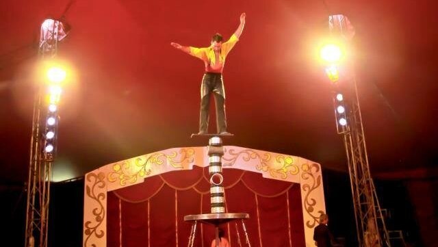 A man balances on a tall stack of pipes inside a red circus tent.