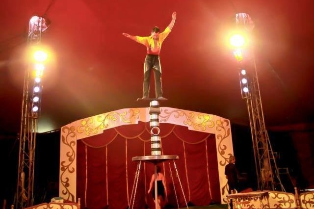 A man balances on a tall stack of pipes inside a red circus tent.